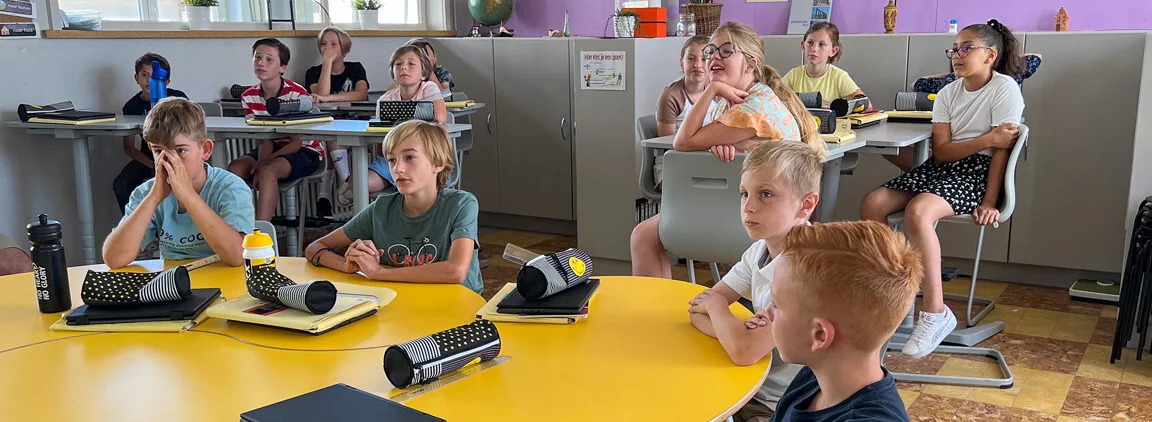 A group of elementary students sit at round tables in a classroom using Learnhub by i3CONNECT, attentively watching the front of the room with tablets and pencil cases on their desks.