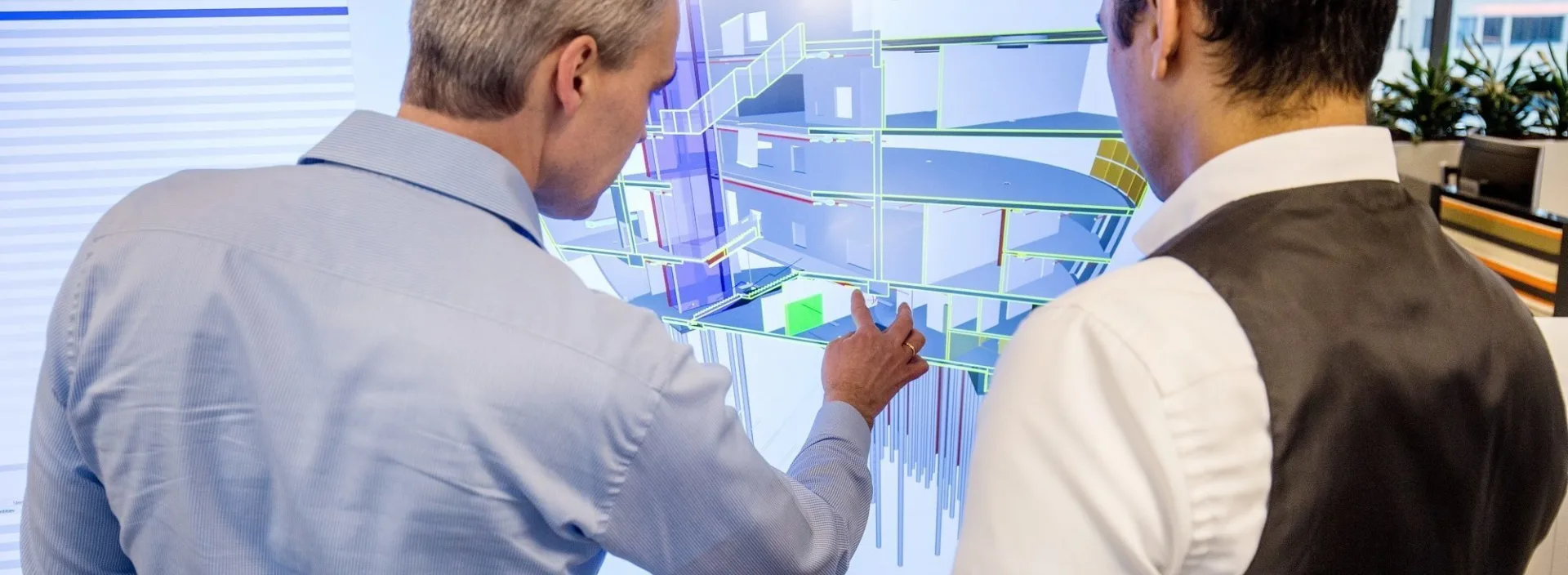 Two men collaborate at a large touchscreen, examining a 3D building model with highlighted structural sections and design details.